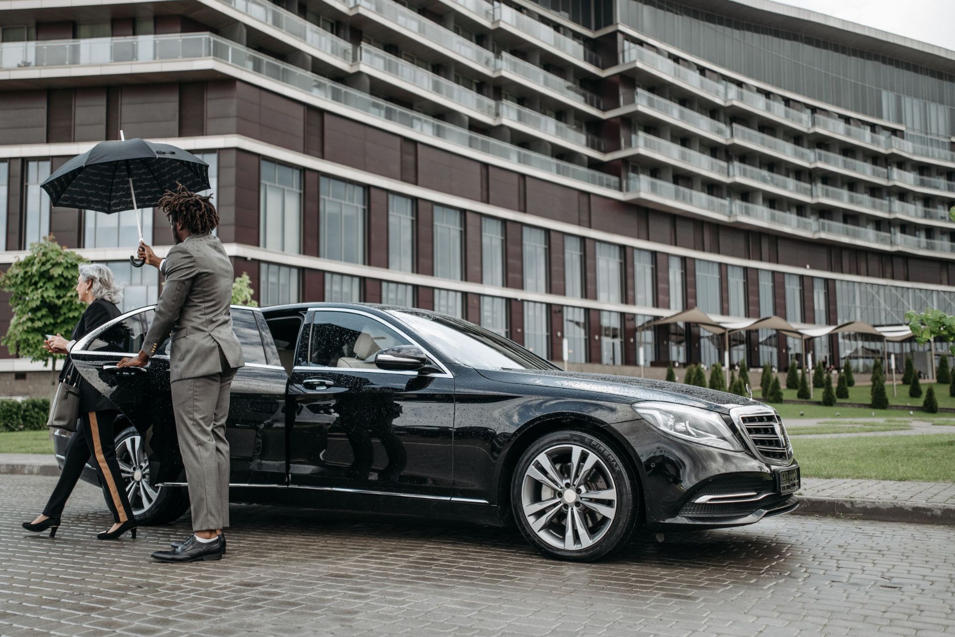 Chauffeur in a tailored suit holding an umbrella and opening the door of a black luxury car for a passenger outside a modern hotel